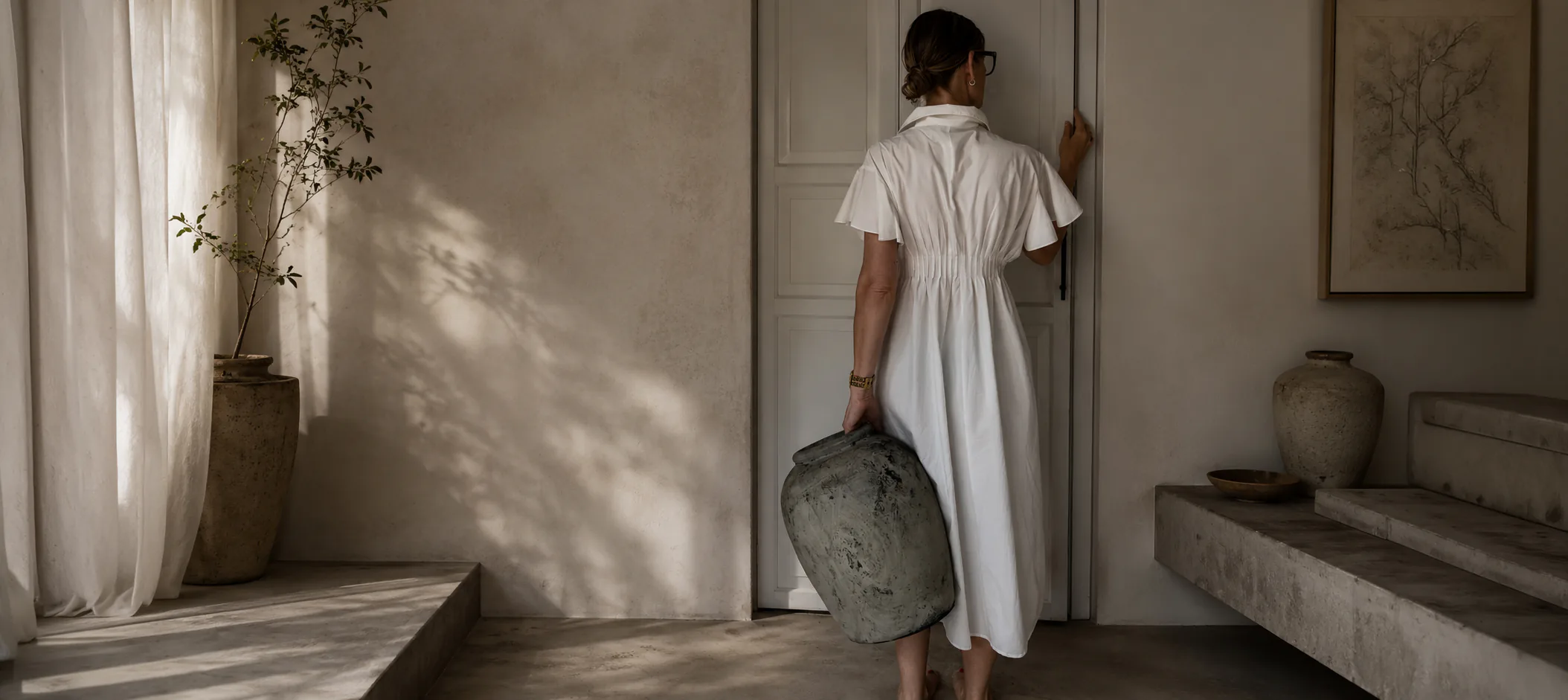 Atelierszene im warmen Tageslicht Person in einem weißen Leinenkleid geht mit einem verwitterten Keramikgefäß durch einen sonnendurchfluteten Türdurchgang